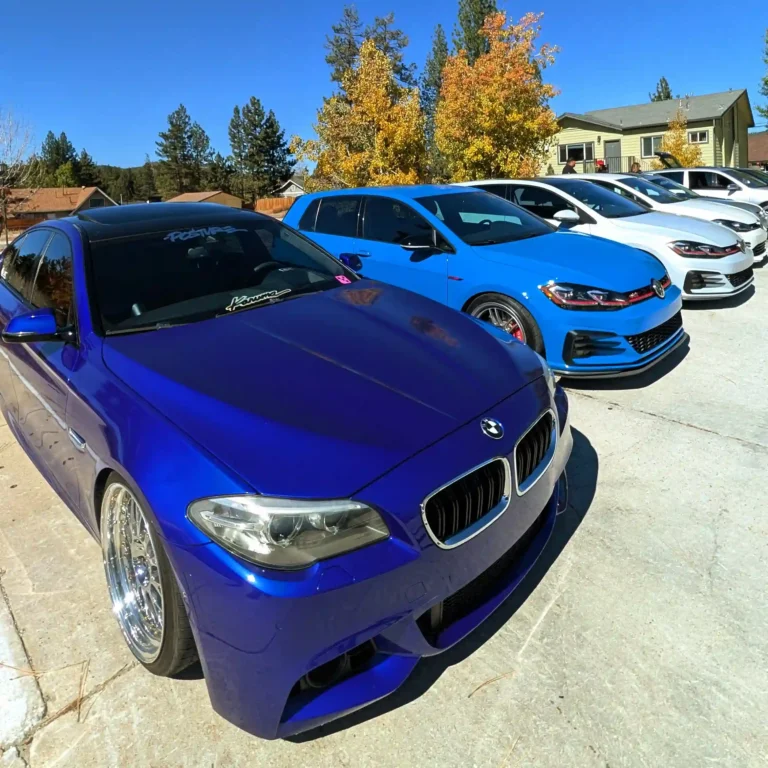 Lineup of European cars including a blue BMW and Volkswagen parked under fall trees at OctoBERGfest in Big Bear Lake