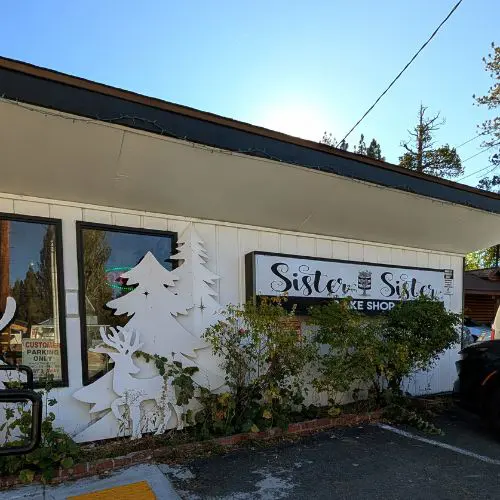 Exterior of Sister My Sister Bake Shop in Big Bear with the main sign partially hidden by bushes and pine trees in the background.