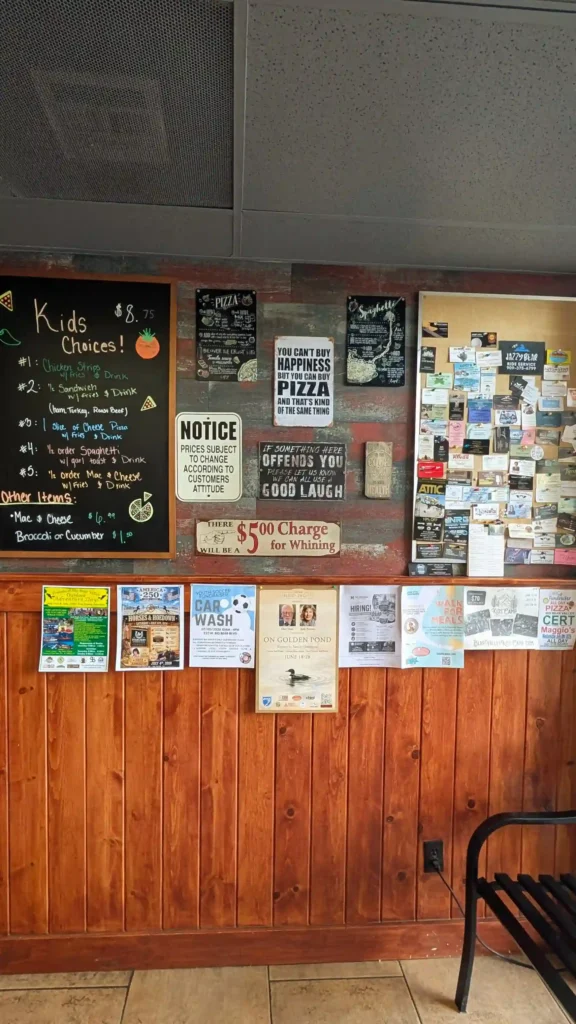 Interior wall at Maggio's Pizza decorated with humorous signs, local event flyers, and rustic wood accents.
